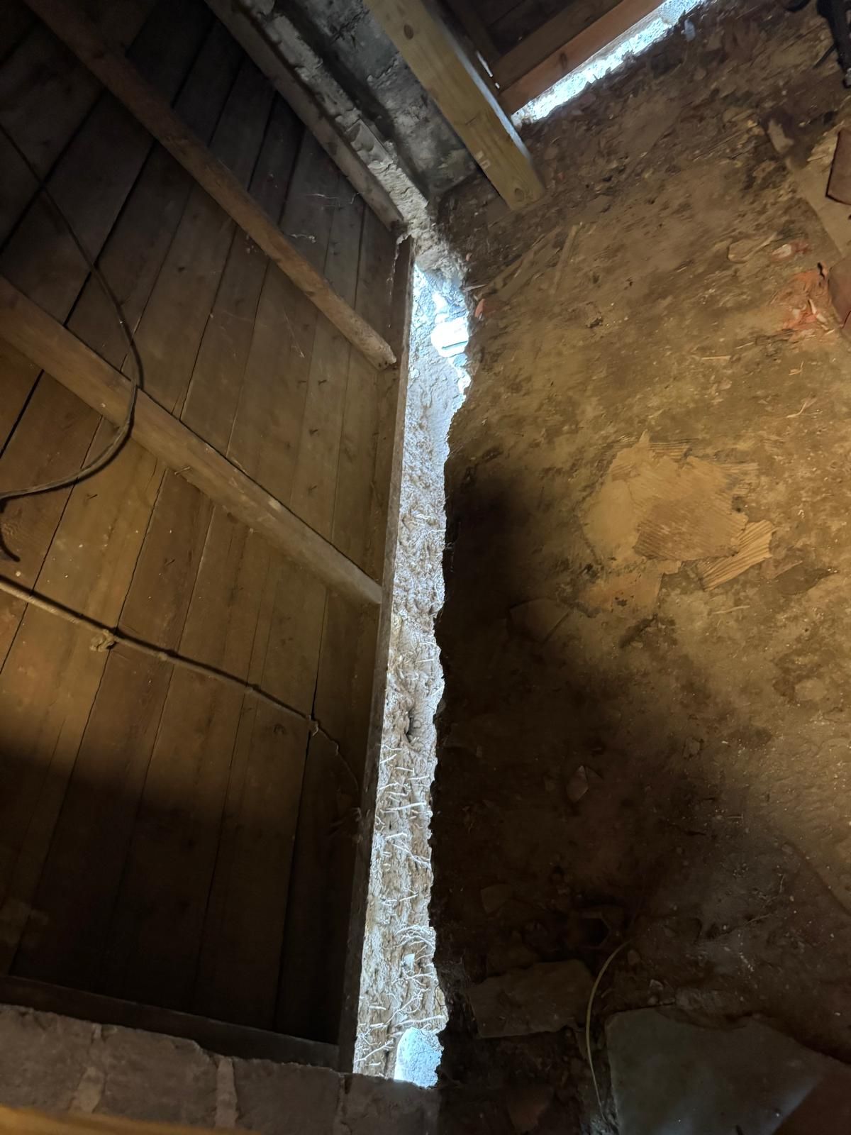 A high-angle view of a gap between a wooden wall and a dirt floor, revealing a layer of white insulation in the crevice.