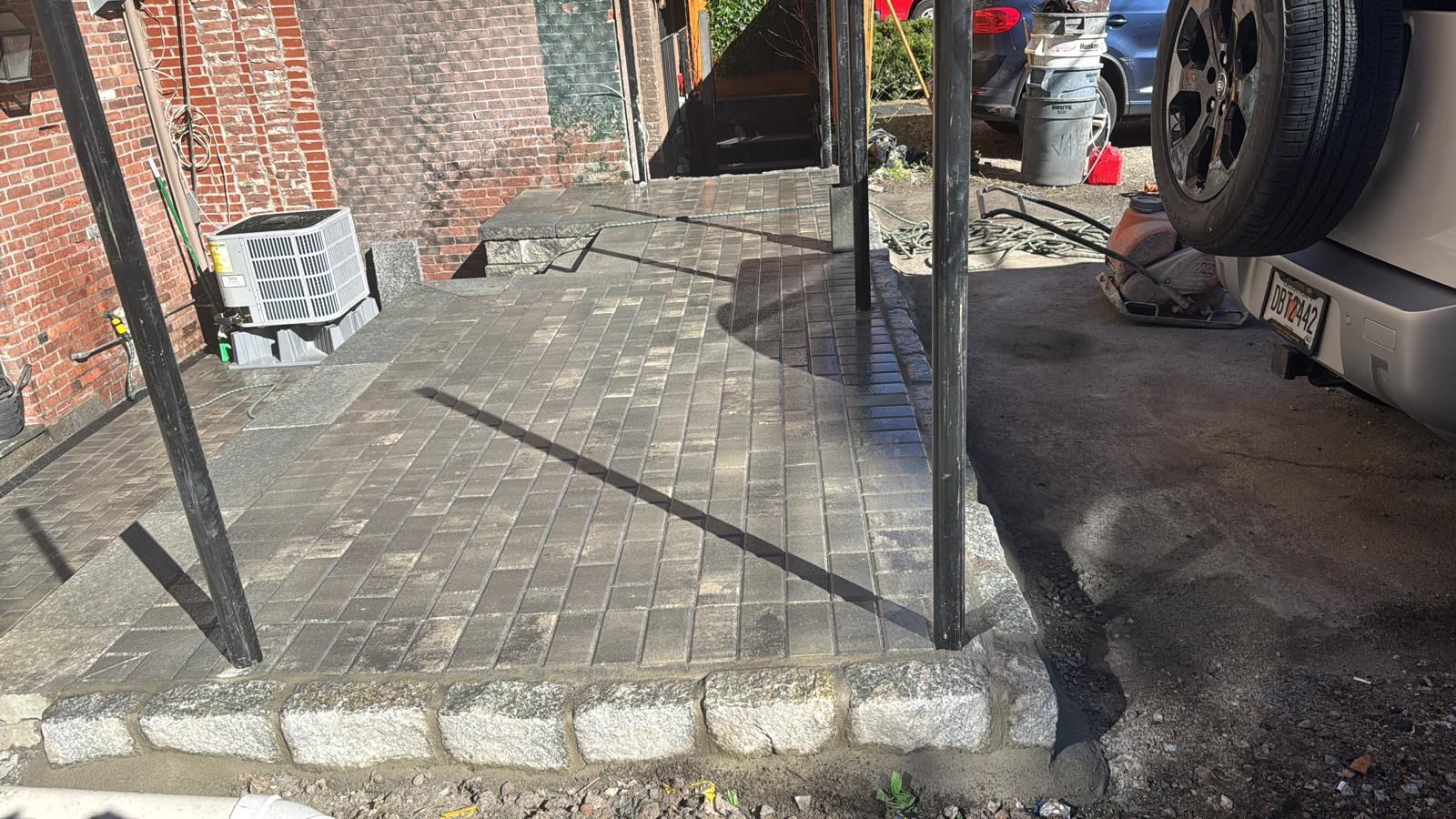 A newly laid grey brick patio with a granite block border, situated in a residential driveway under a metal carport.