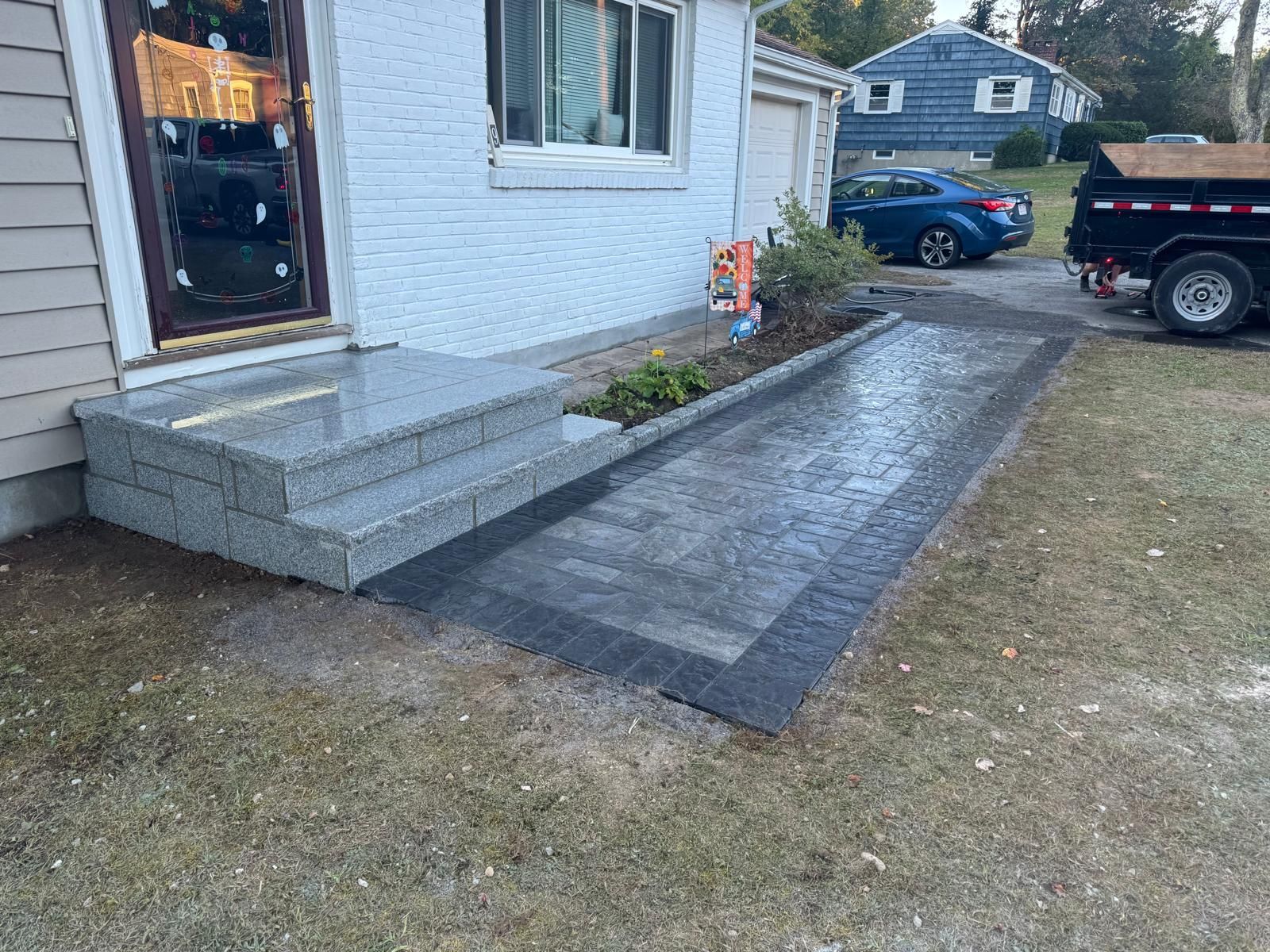 Newly installed stone steps leading to a house door next to a gray paver walkway, with a driveway and car in the background.