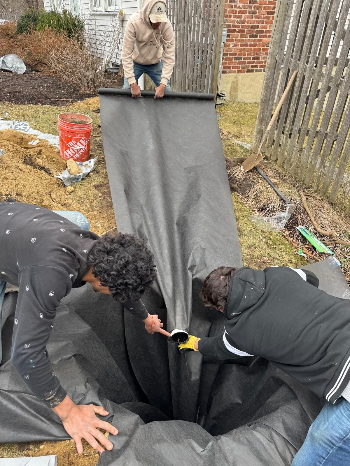 Three people work together to line a deep, excavated trench with black landscape fabric in a residential yard.