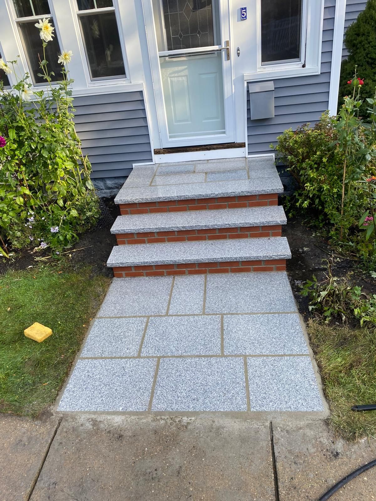 A new gray granite entryway with three steps leading to a front door, flanked by greenery and brick accents.