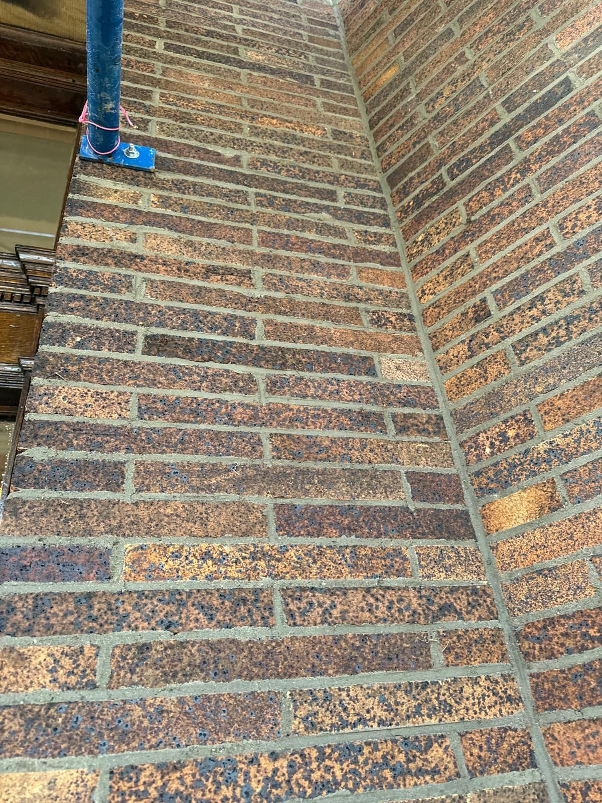 A corner of a brown brick wall with a blue metal scaffolding post secured to the floor at the edge.
