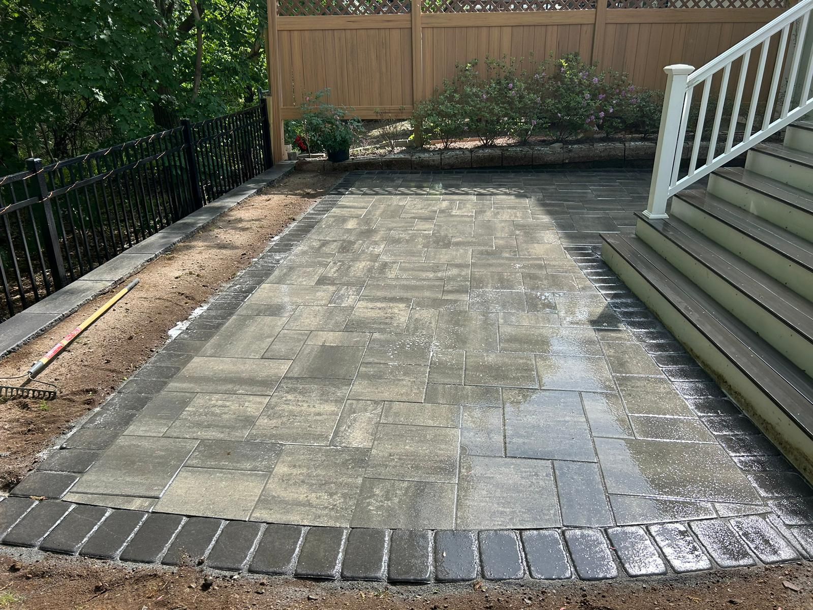 A newly installed grey paver patio with a dark border, situated next to wooden stairs and a privacy fence.