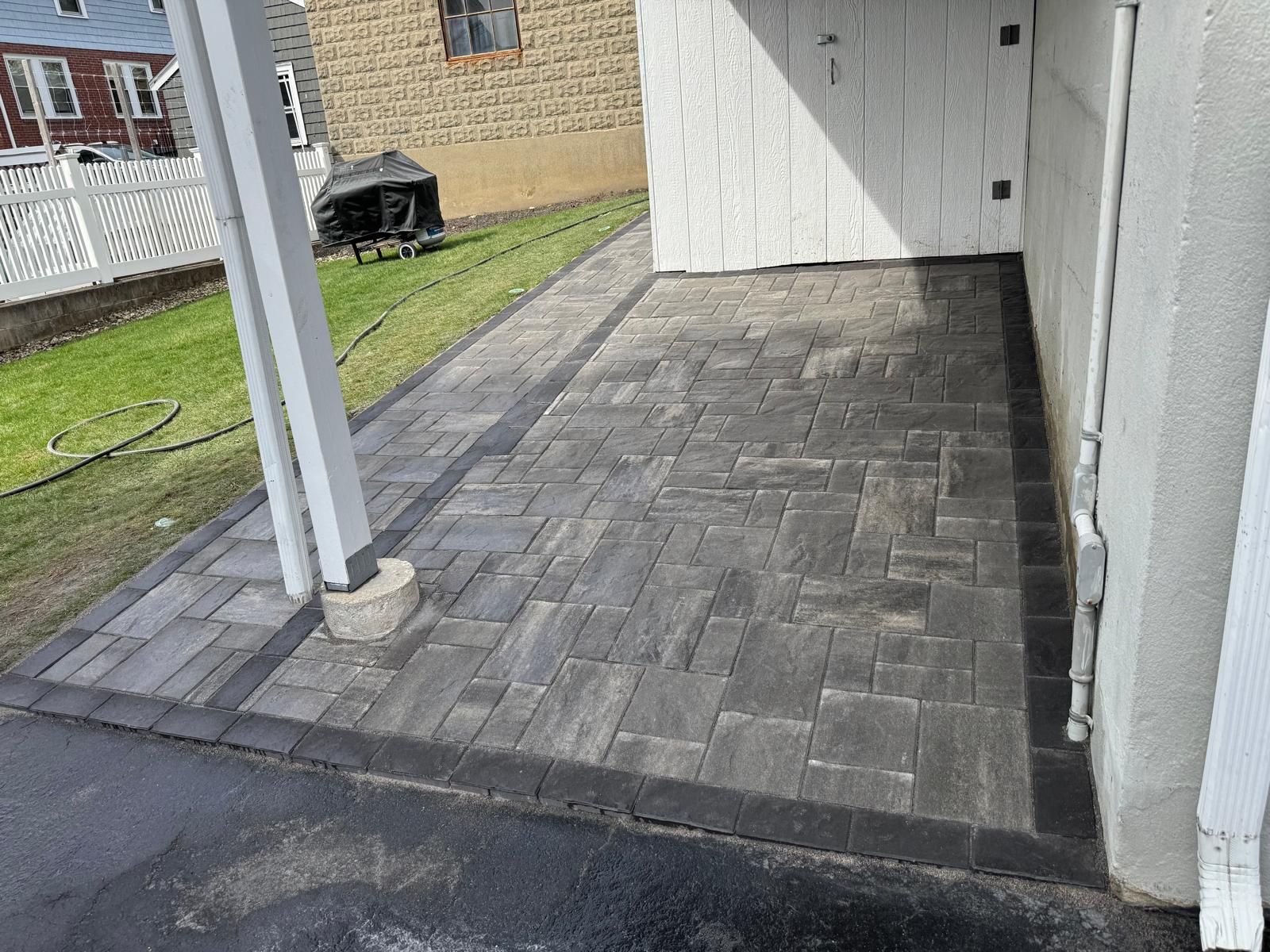 A paved patio with grey rectangular stones, a dark border, and a white support post next to a building and grass.