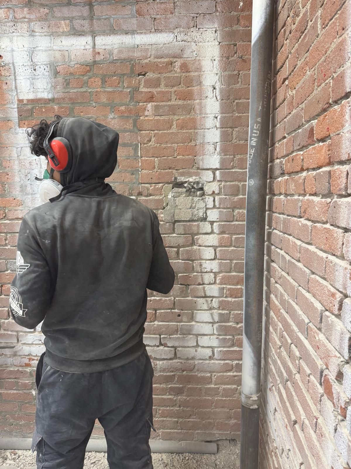 A person wearing protective gear and a dark hoodie faces a weathered brick wall while working on a construction site.