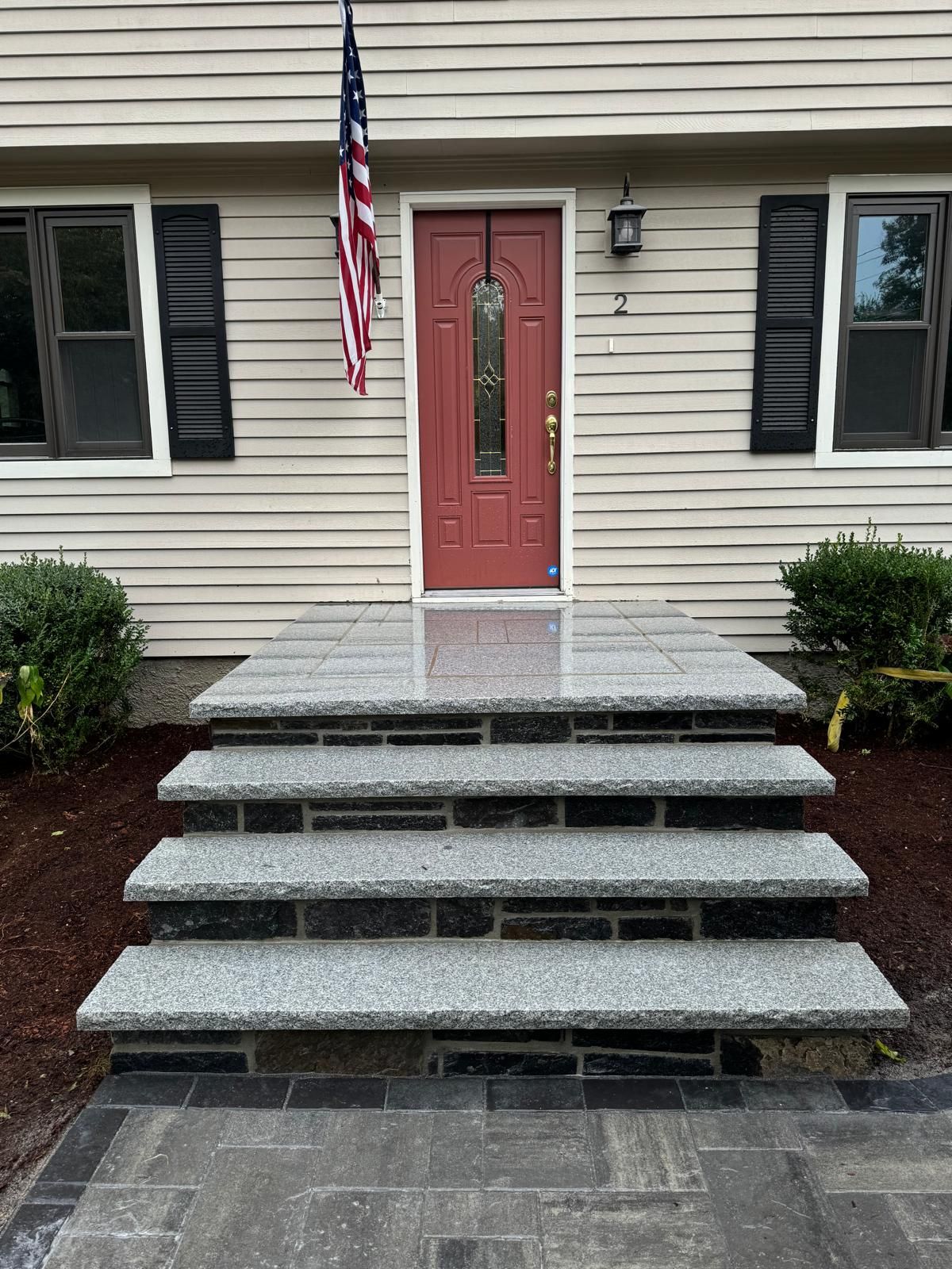 A red front door with a flag above it, accessed by four gray granite steps leading up to a landing in front of the home.