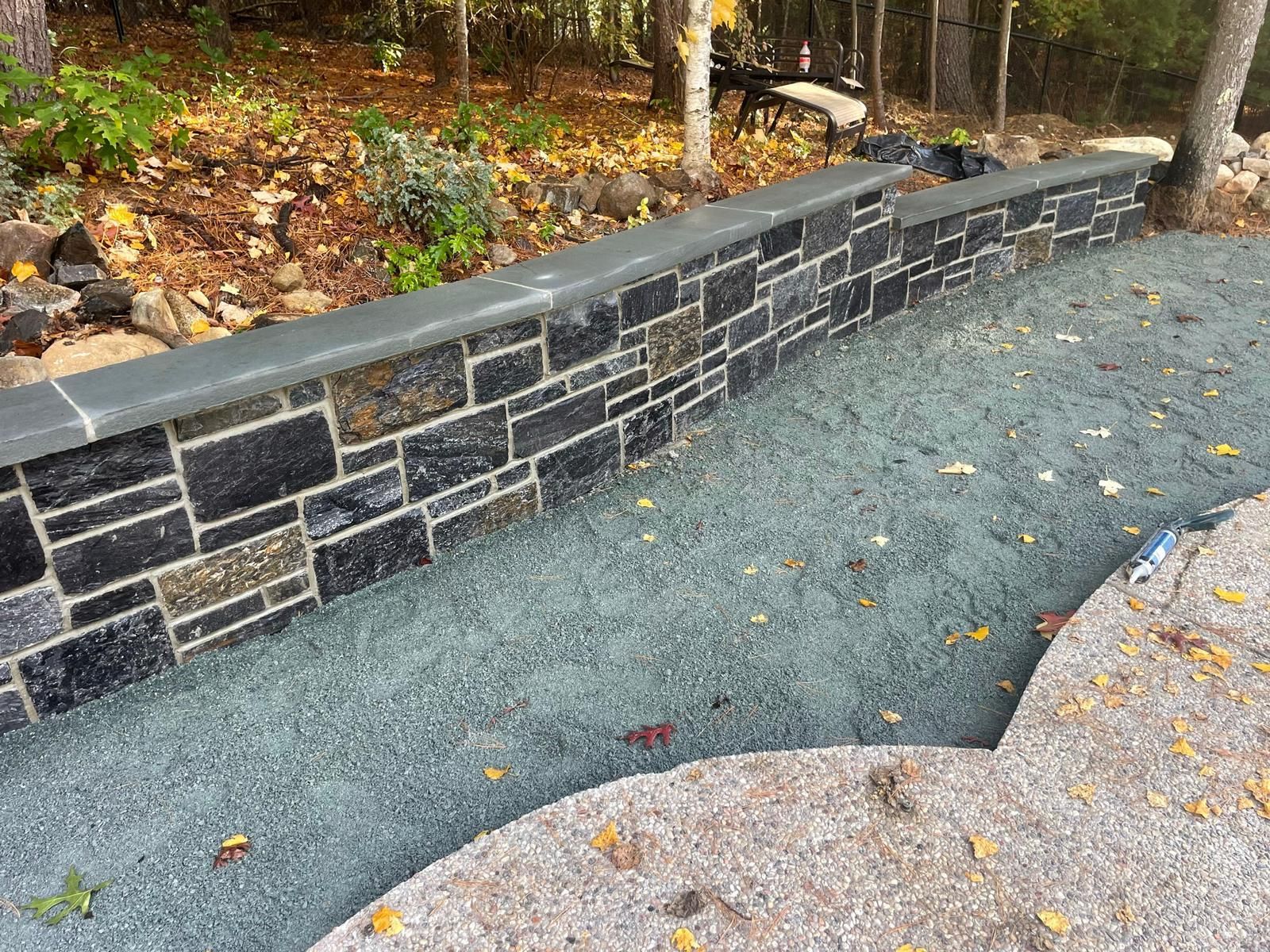 A stone retaining wall with a smooth gray cap, bordering a gravel path and a woodsy landscape with scattered autumn leaves.