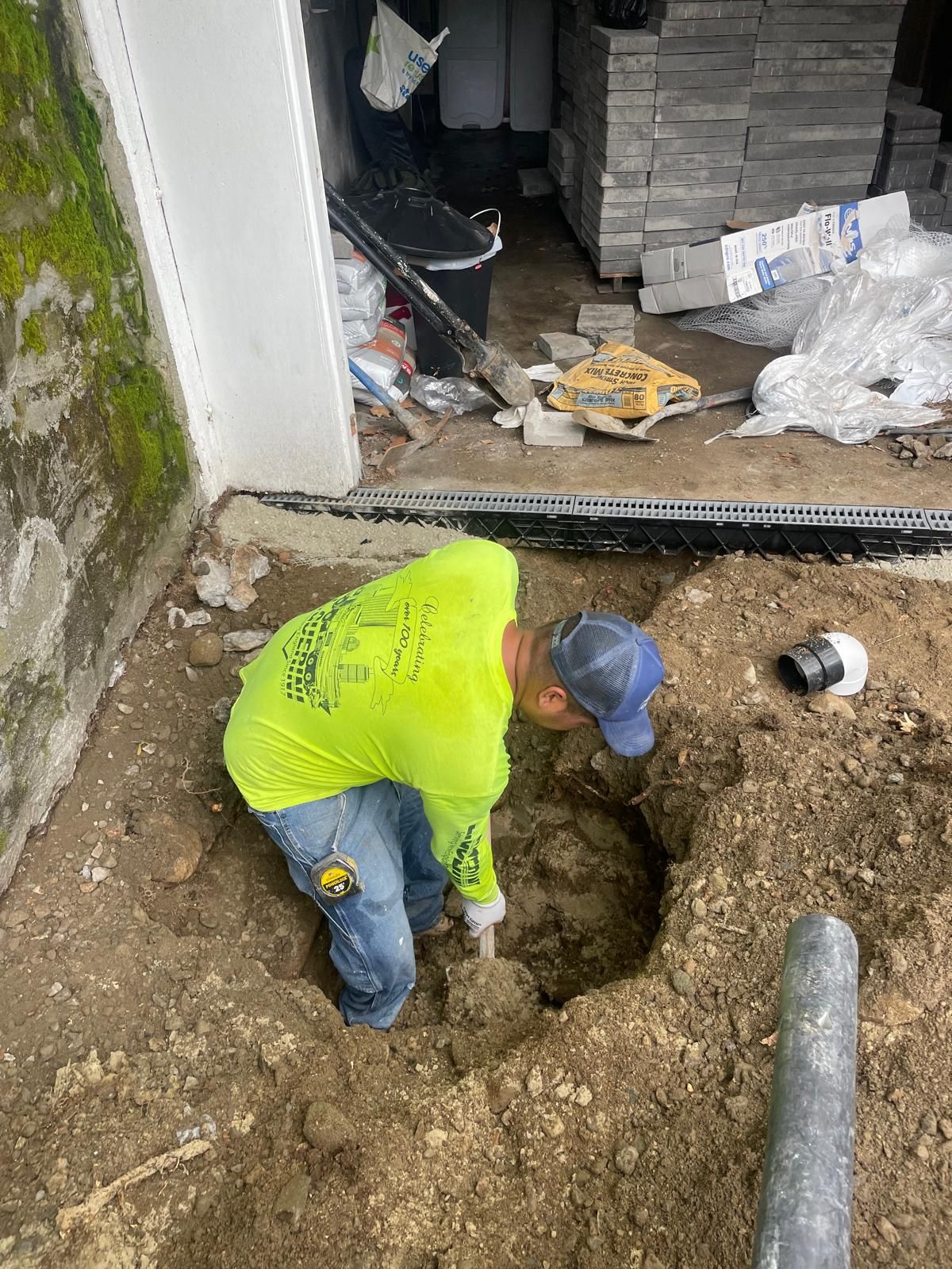 A person in a neon yellow shirt and a blue cap works in an excavated dirt hole next to a wall.