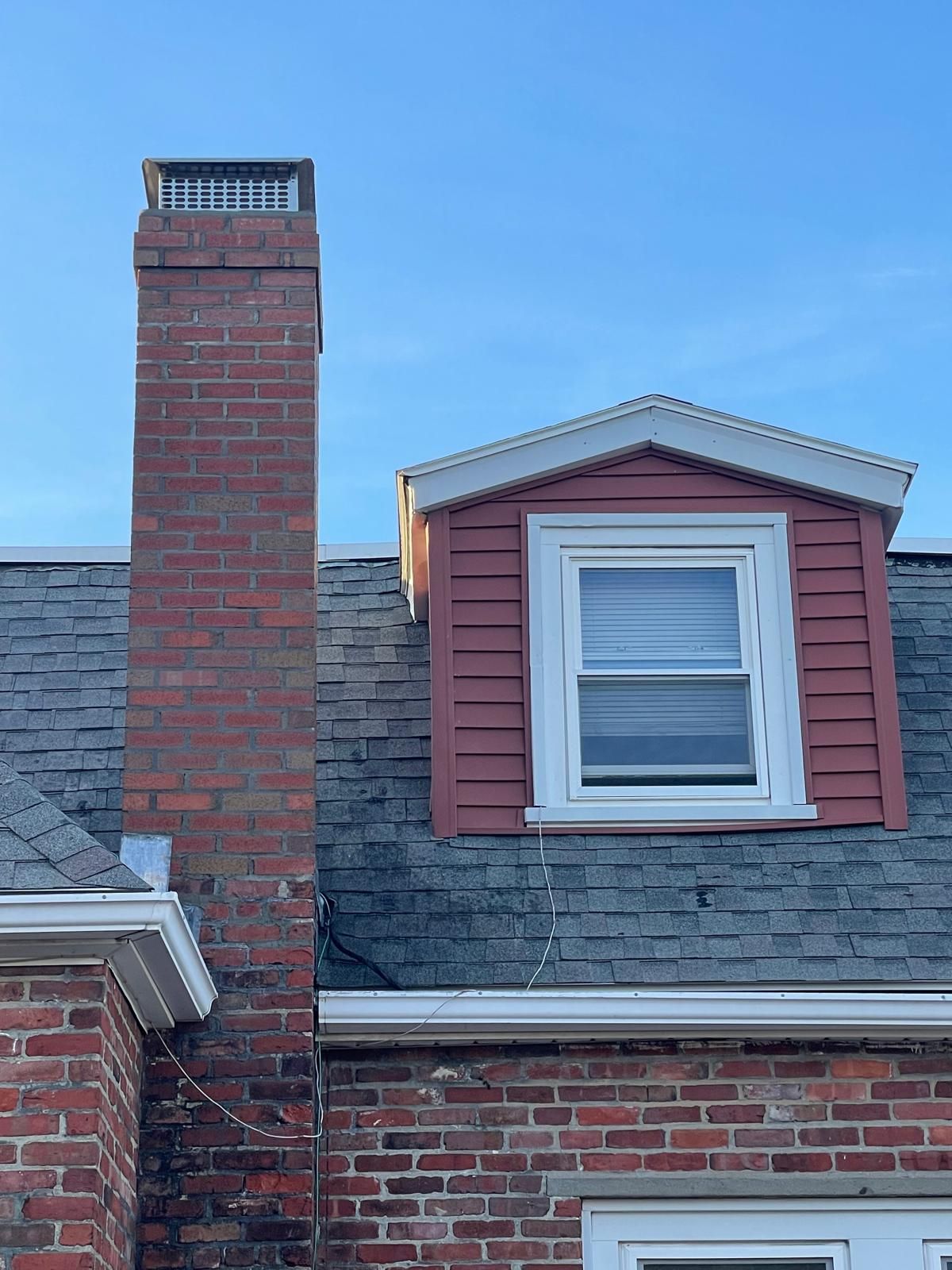 A red brick chimney topped with a metal flue, adjacent to a dormer window with red siding and a white frame on a gray roof.