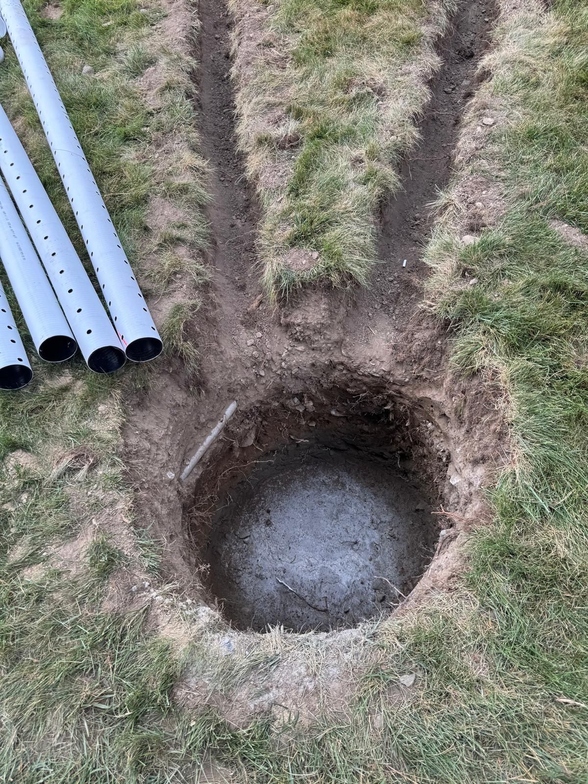 A deep circular hole dug into grass, with two narrow trenches extending outward and four perforated metal poles to the side.