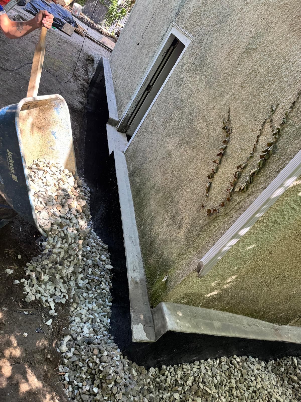 A wheelbarrow pours gravel into a trench lined with a black waterproof membrane along the exterior wall of a building.