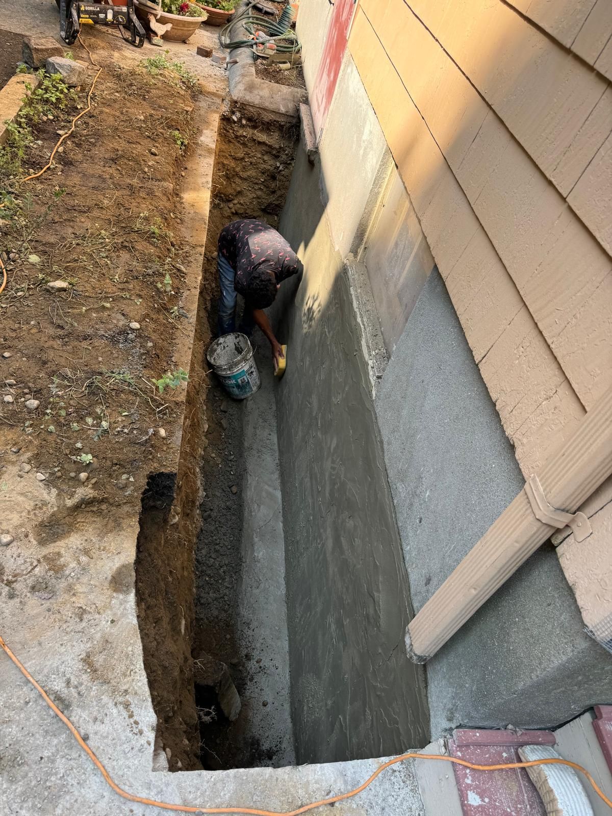 A worker applies waterproof sealant to the foundation of a house exterior in an excavated trench.