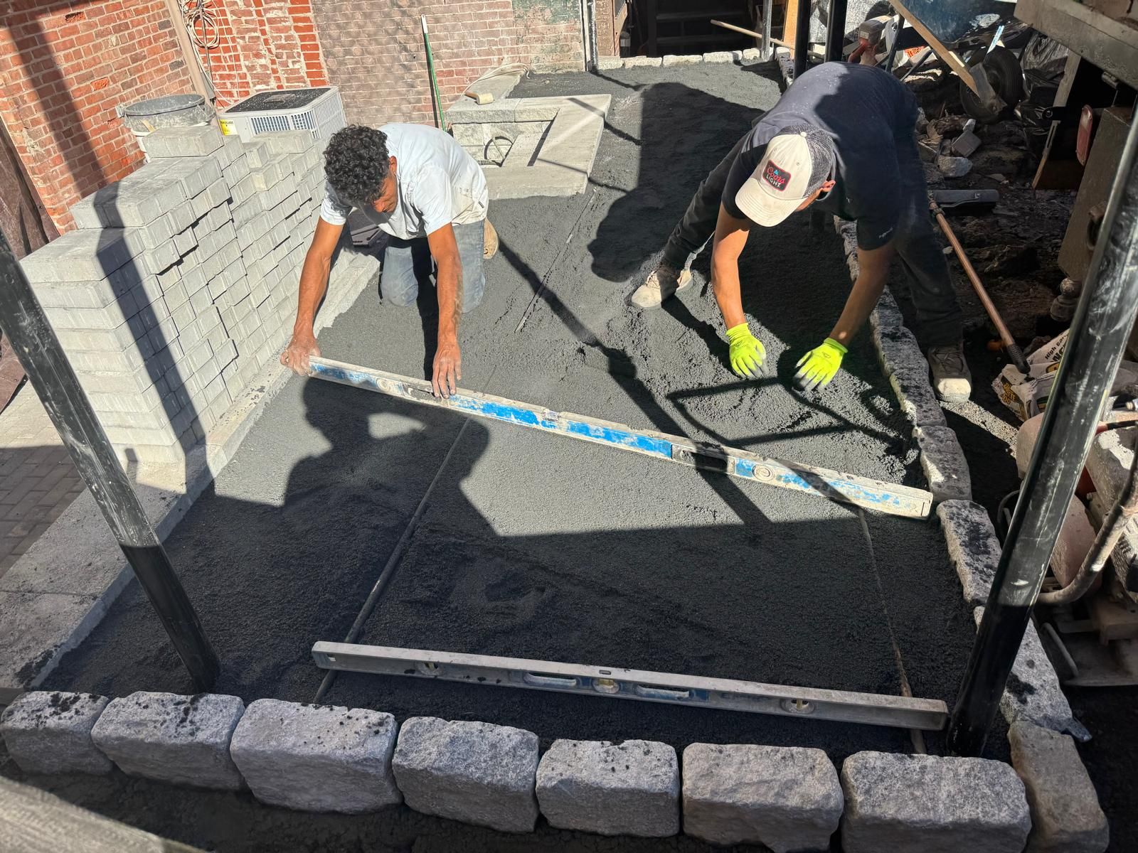 Two workers use metal levels to smooth gravel bedding for a stone patio project.