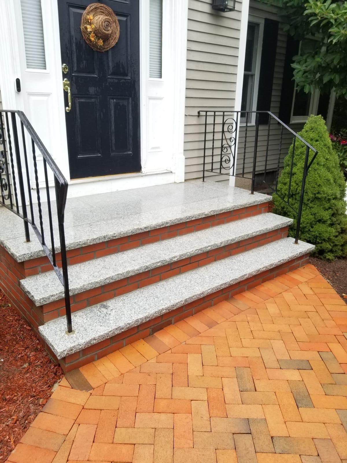 Front steps with light gray granite treads, brick risers, and black metal railings leading to a black front door.
