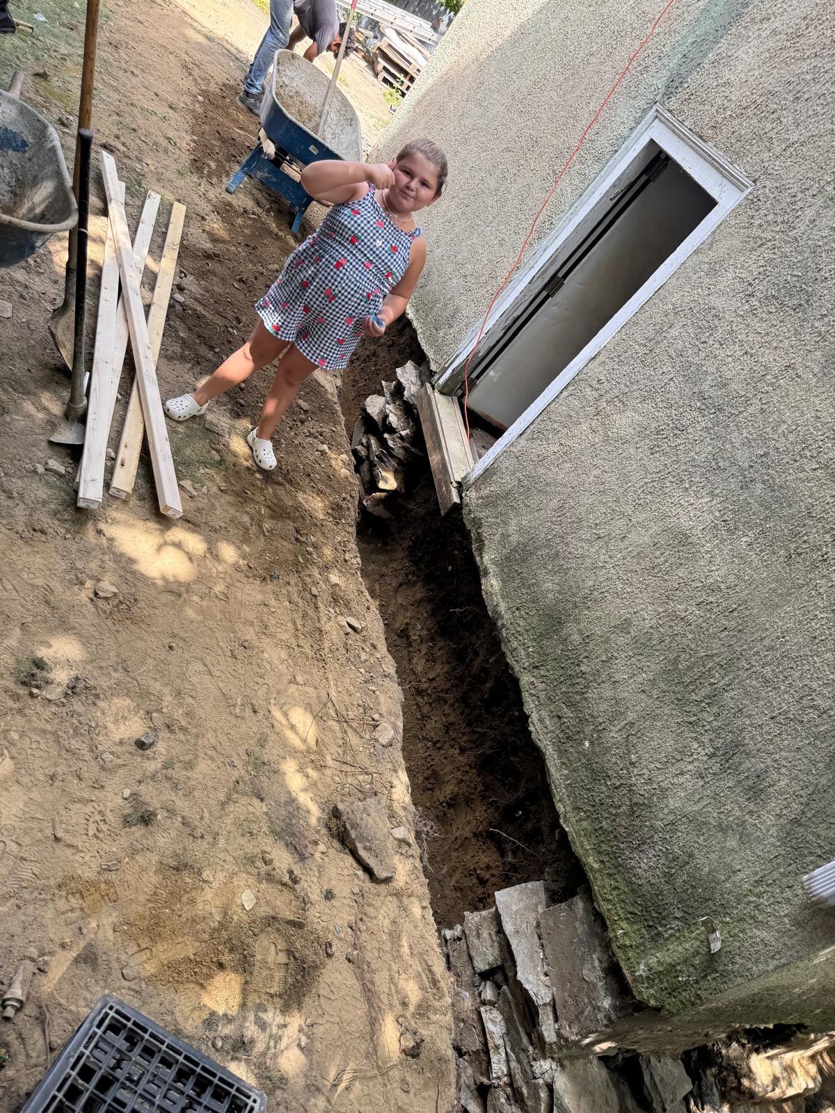 A young person stands next to a deep trench dug along the side of a building foundation, with construction tools nearby.