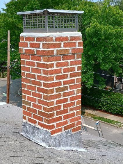 A rectangular brick chimney on a shingled roof, featuring a metal cap and flashing at the base against the roof shingles.