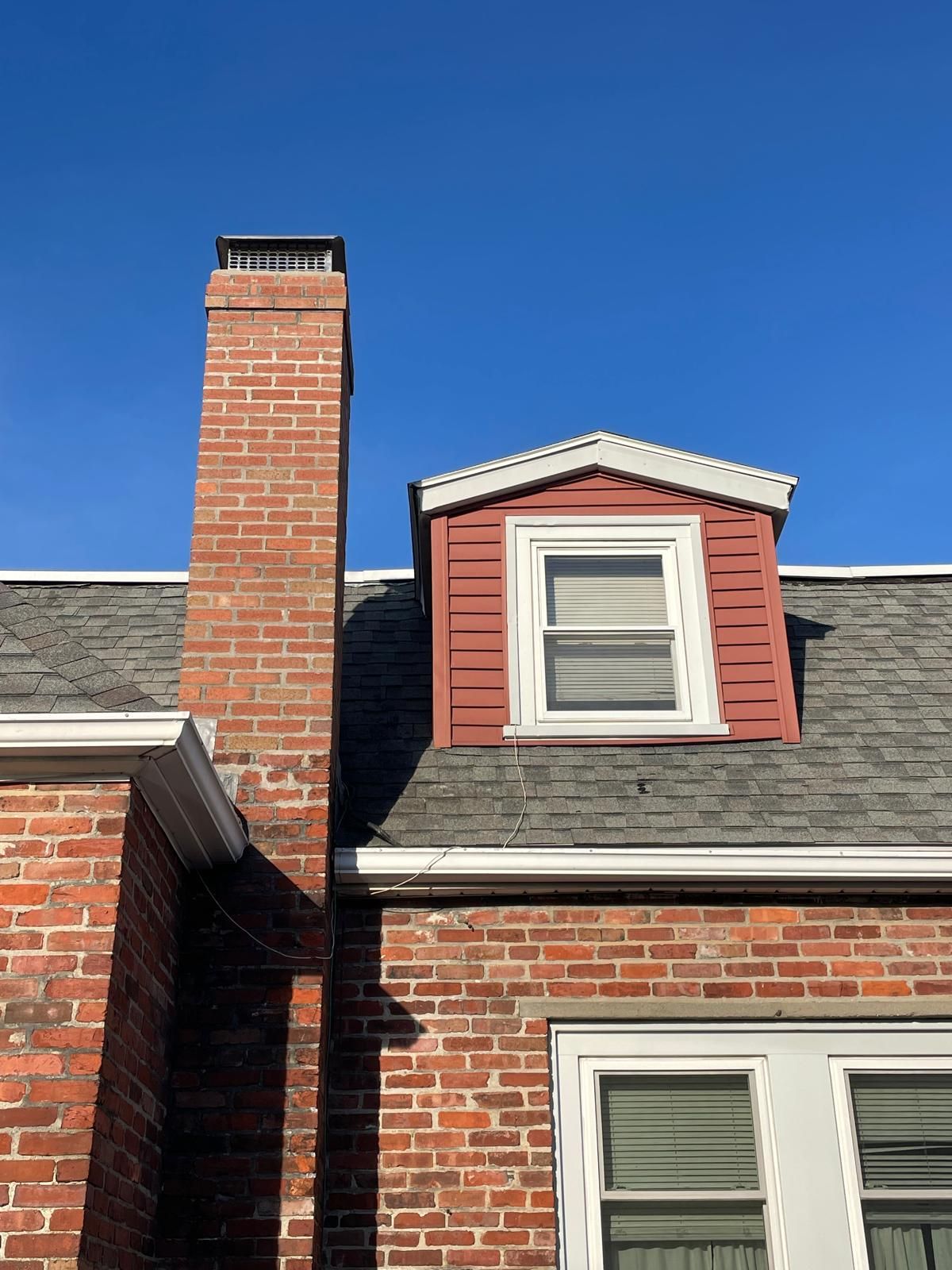 A red brick chimney rises next to a small dormer window with red siding on a house roof under a clear blue sky.
