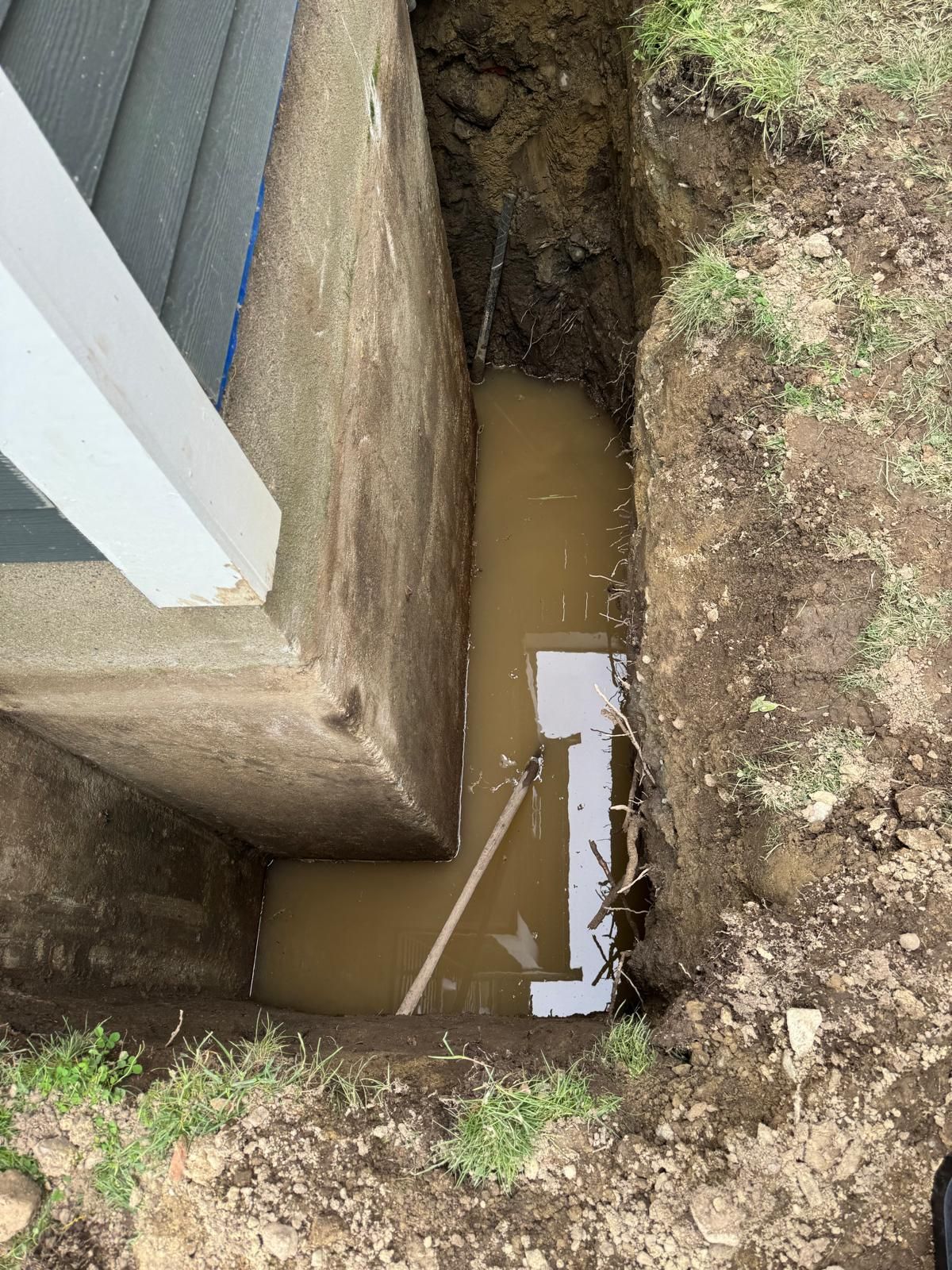 A deep trench dug beside a house foundation, partially filled with muddy water and exposing a pipe section.