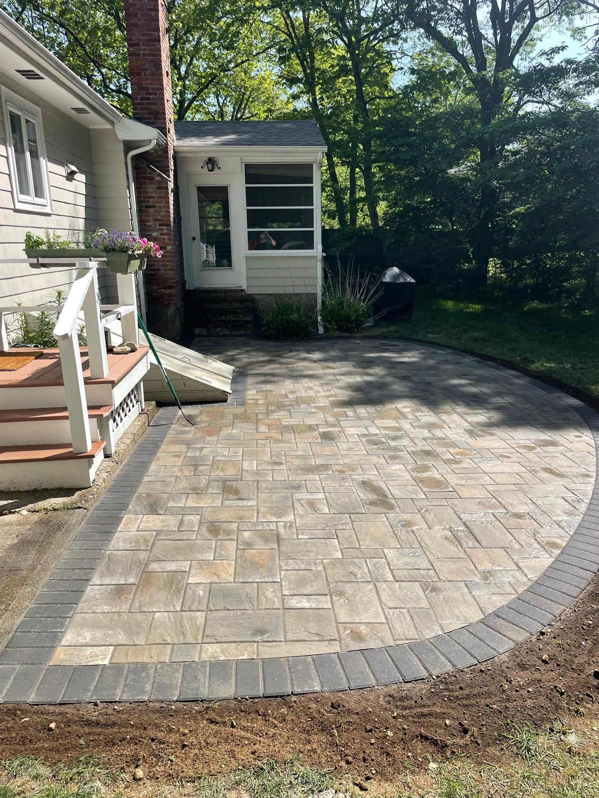 A newly installed rectangular paver patio with a curved border, located beside a house with white siding and a chimney.