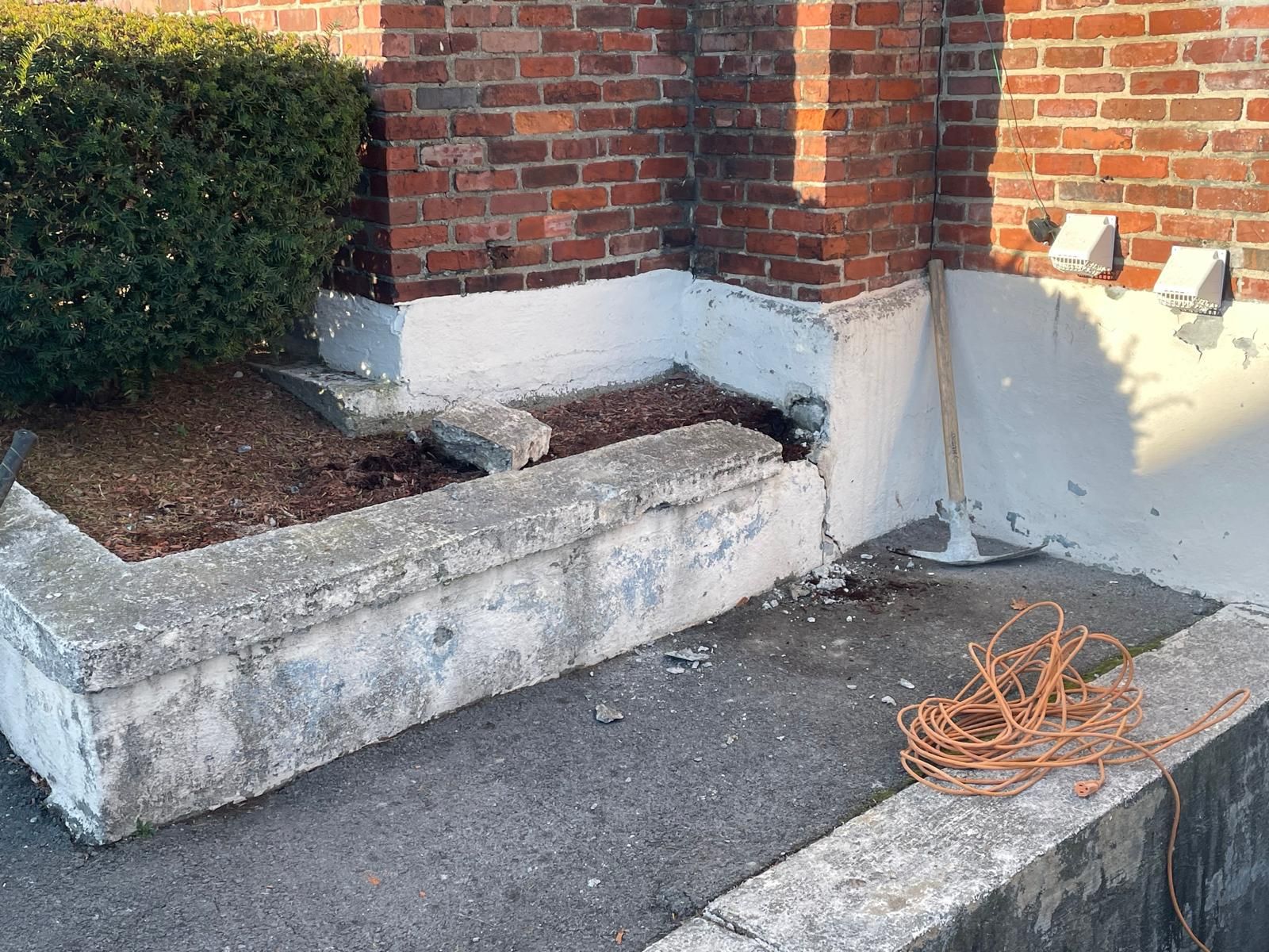 A brick building corner with a damaged concrete planter, a coiled orange extension cord, and a small gardening tool.