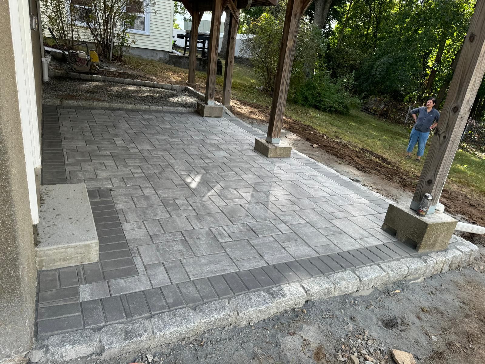 A newly installed grey paver patio with a dark border under a wooden pavilion, with a person standing in the background.