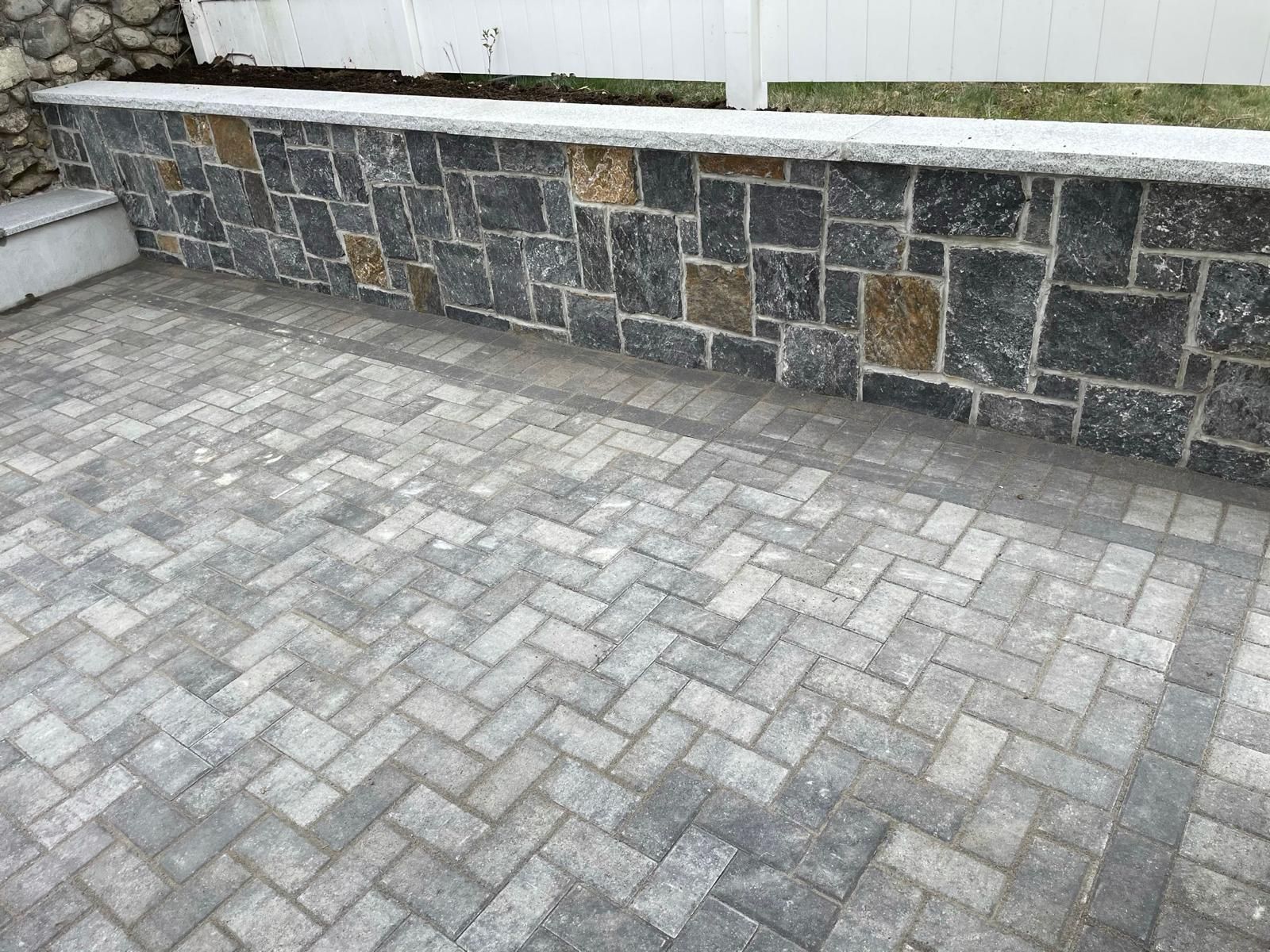 A gray stone paver patio in a herringbone pattern sits below a low retaining wall made of dark, multi-sized stone blocks.