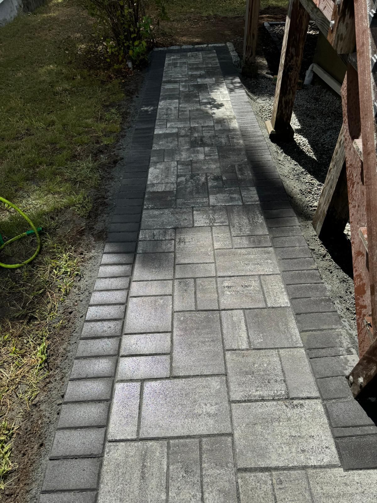 A gray stone paver walkway with a dark border, leading through an outdoor area with a ladder visible on the right.