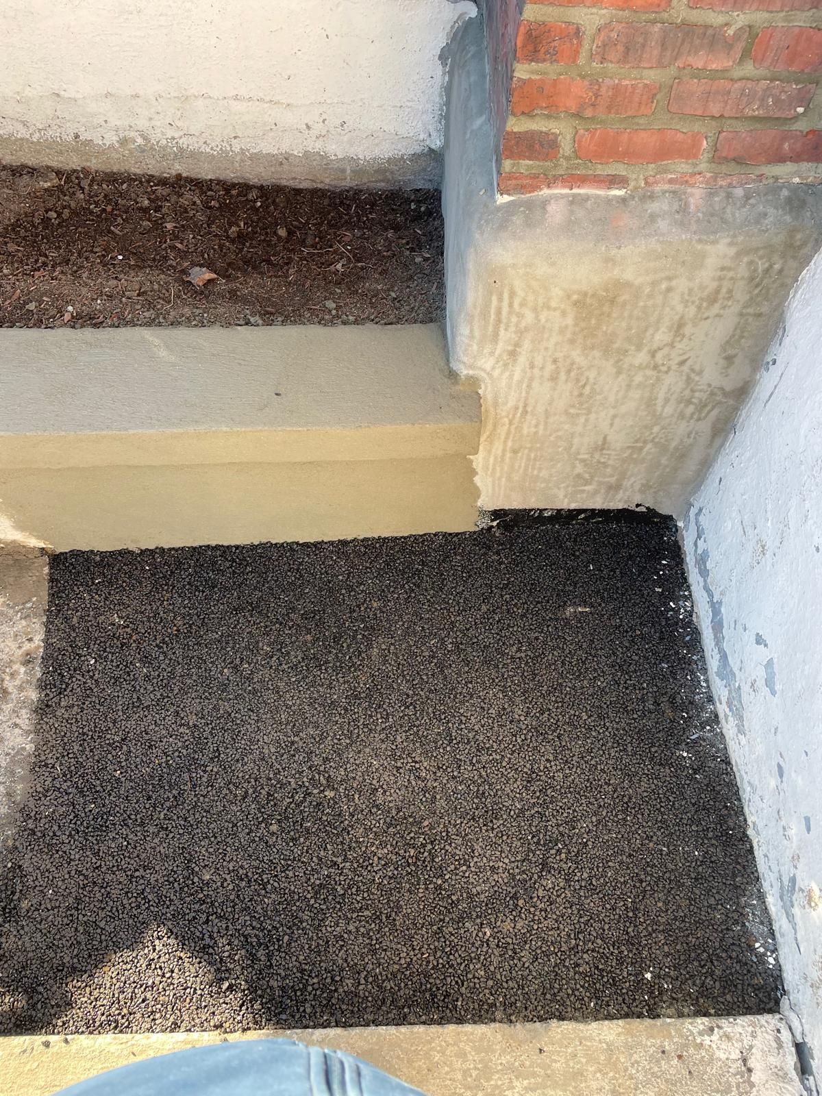 A textured black doormat placed at the base of a concrete step near a brick and stucco building wall.