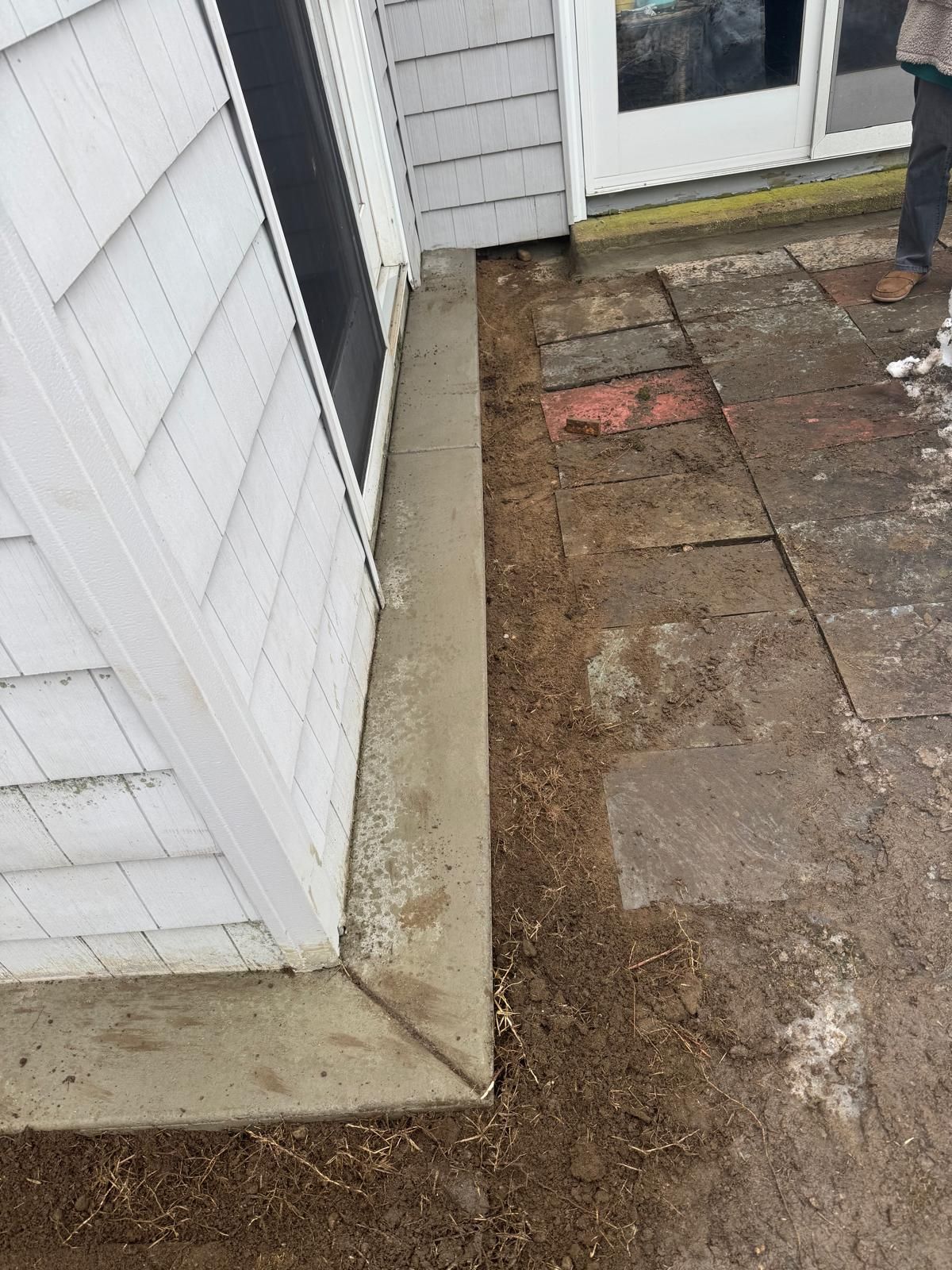 A view of a corner of a white shingled house showing a newly poured concrete foundation beside a dirt patio area.