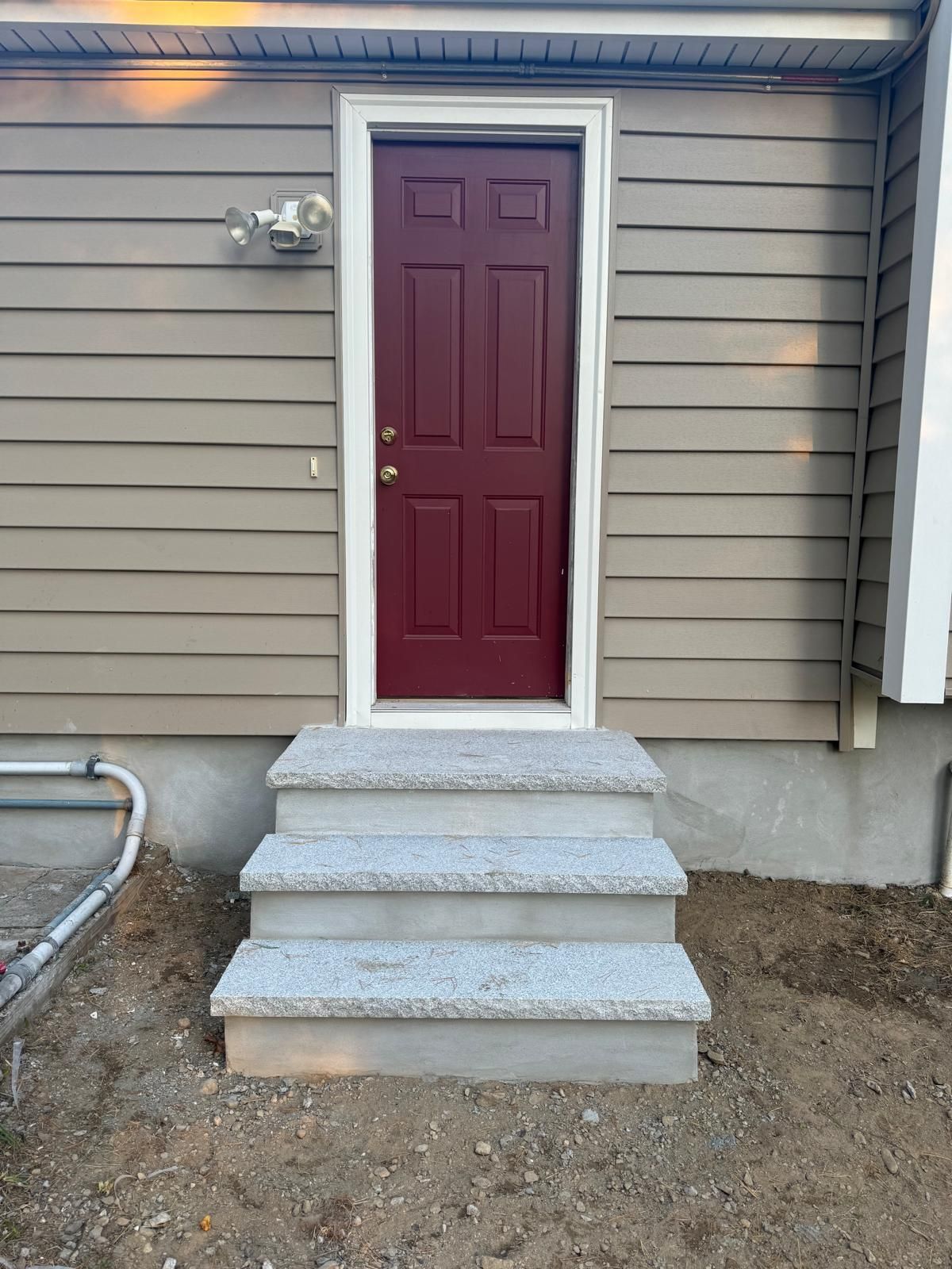 A red door with a white frame above three light gray stone steps against tan horizontal siding.