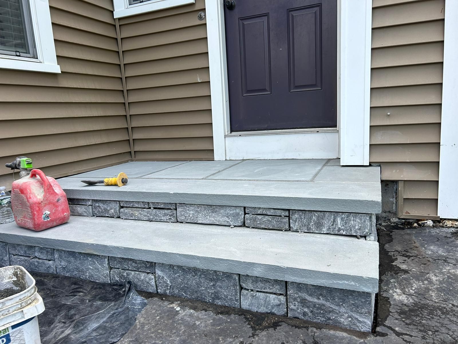 New stone porch steps leading to a dark brown door on a house with tan siding, with a red fuel container nearby.