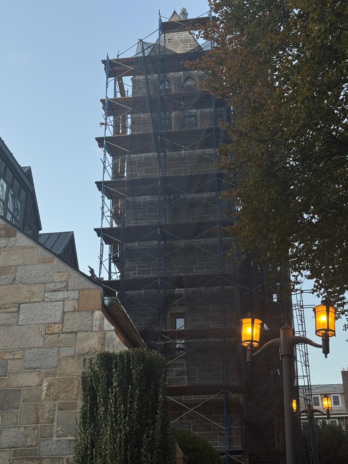 A tall, stone clock tower encased in scaffolding stands under a clear blue sky, next to a stone building and street lamps.