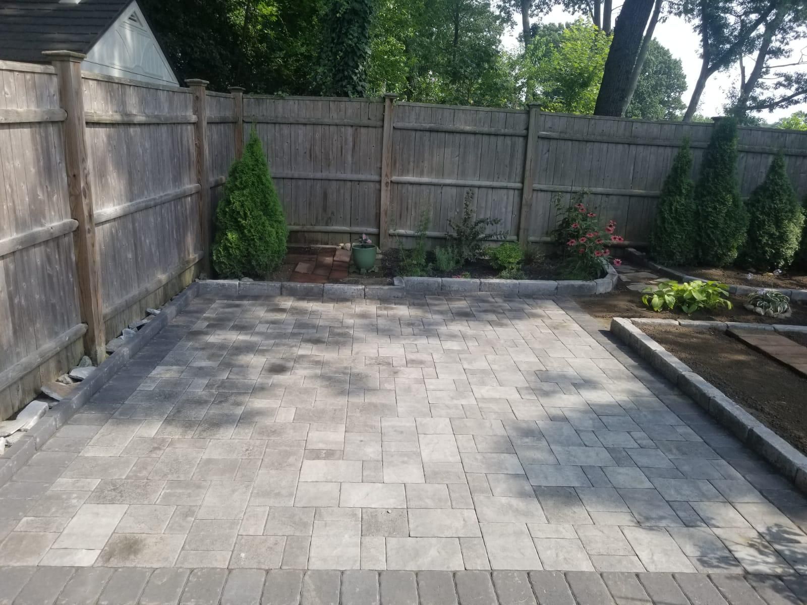A gray stone patio with a dark border, situated in a backyard next to a wooden fence and landscaped garden beds.