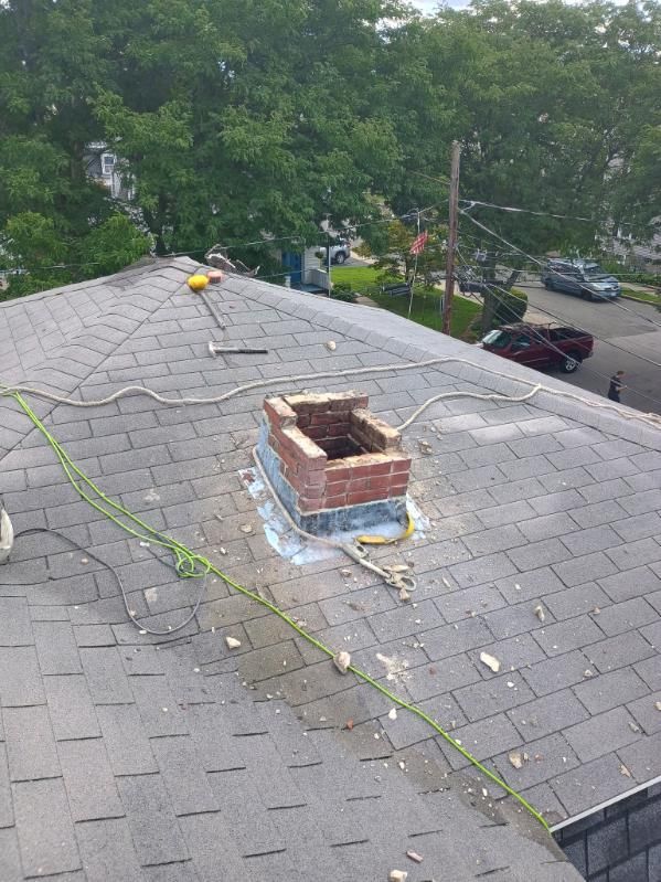 An aerial view of a gray asphalt shingle roof featuring a partially demolished brick chimney with exposed flashing.
