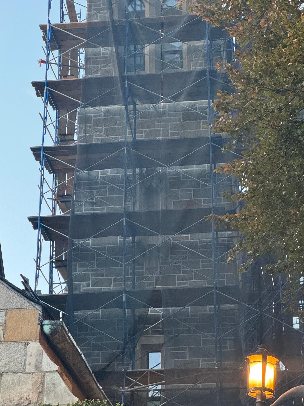 A stone church tower under renovation, surrounded by blue scaffolding draped with black safety netting.