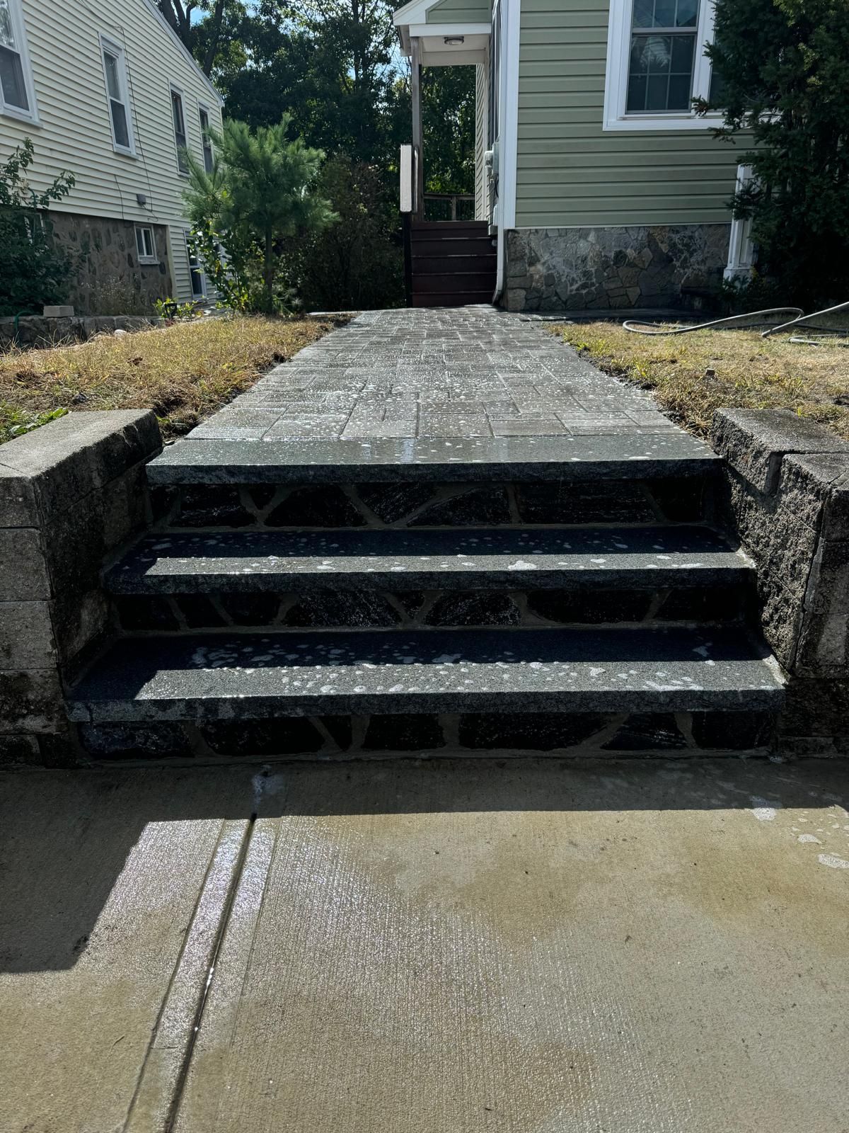 A concrete walkway leading to three dark stone steps with stone retaining walls, leading to a house entrance.