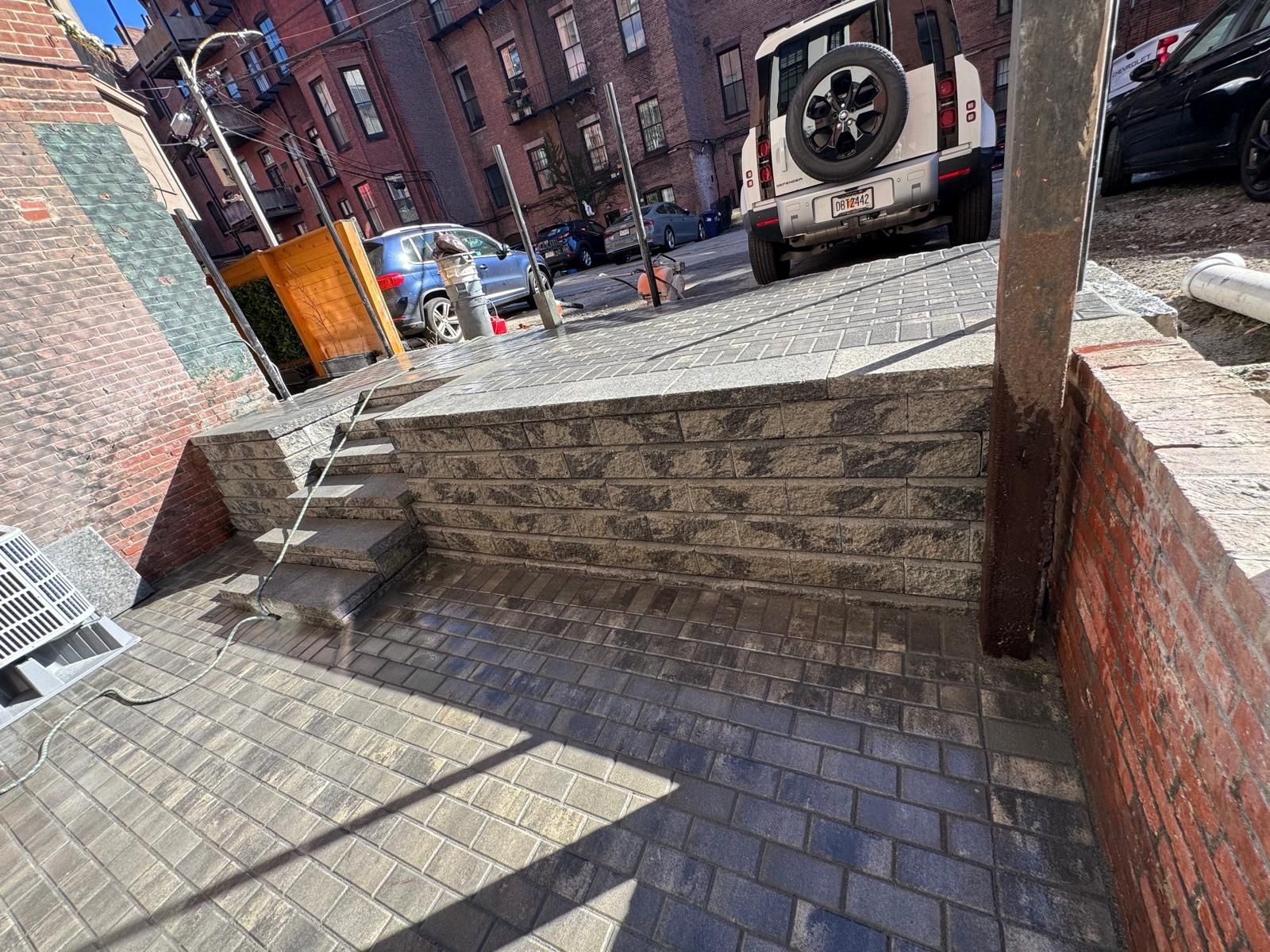 A paved alleyway featuring a raised stone wall with small concrete steps leading to a parking area with parked vehicles.