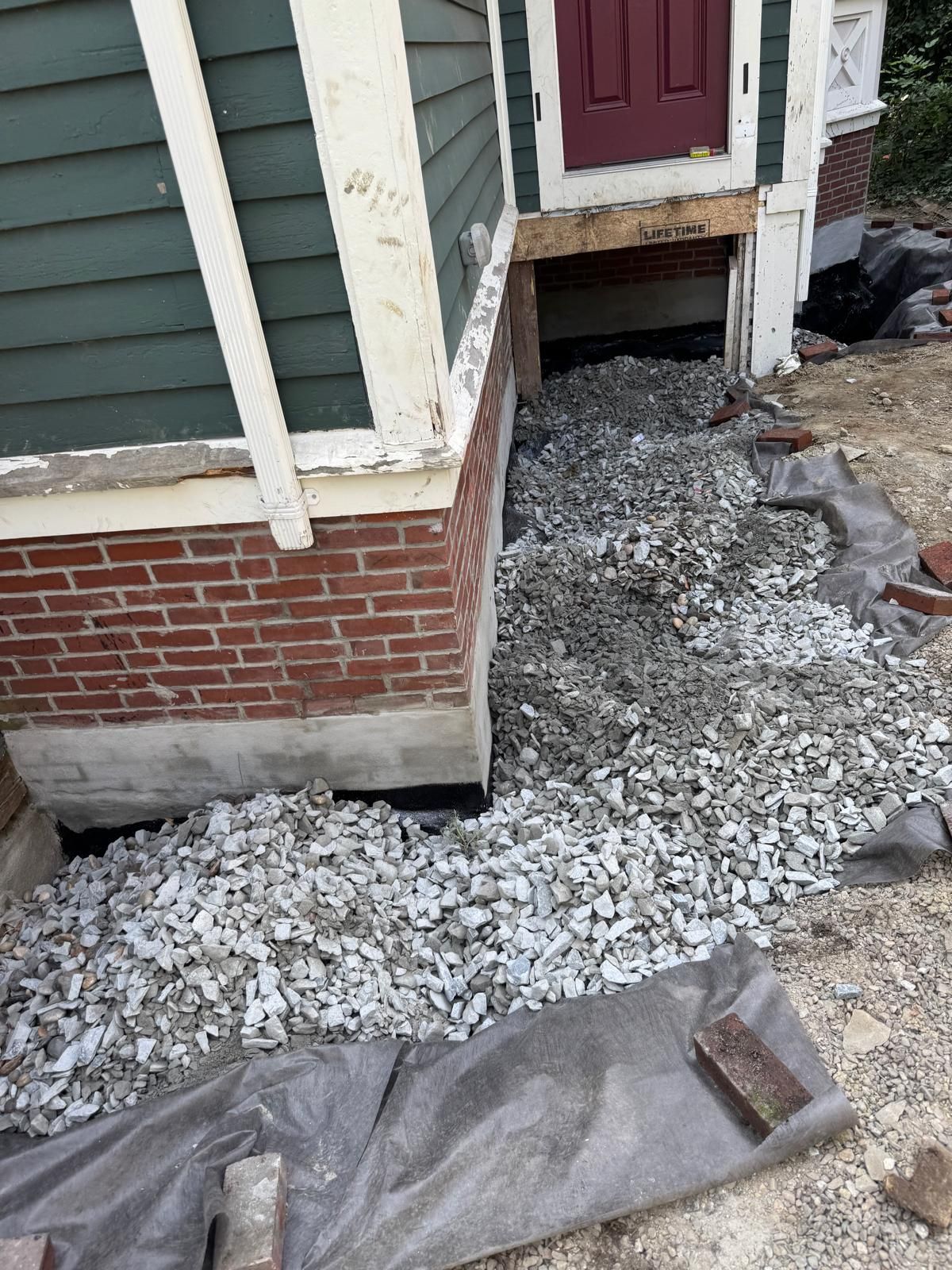 Exterior view of a house foundation under construction, with gravel laid over ground fabric against brick and siding walls.