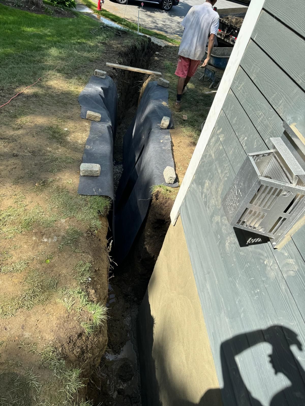 A person walks beside a deep trench dug along a house foundation, lined with black landscape fabric held down by bricks.