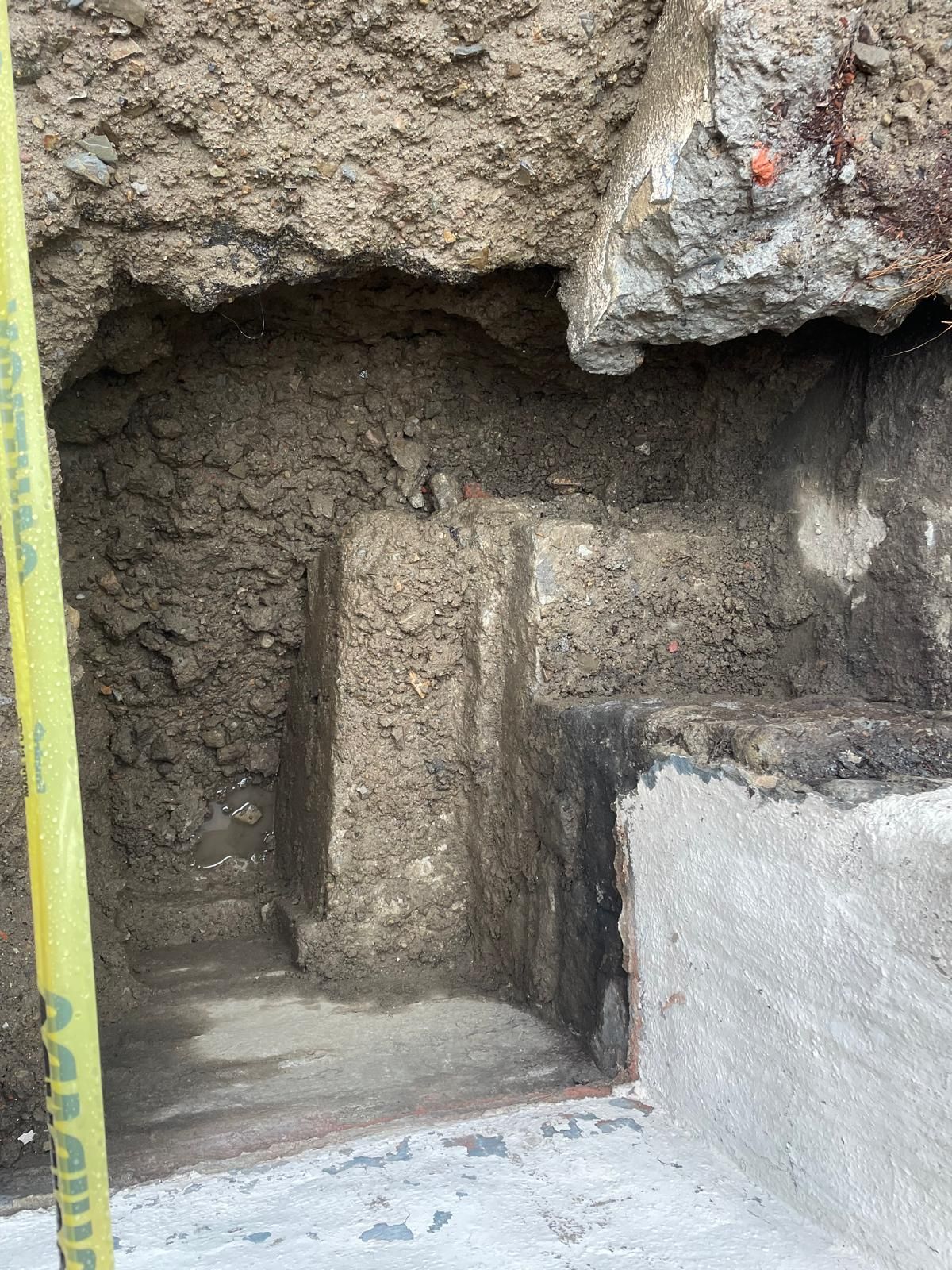 A deep, L-shaped excavation pit in dirt next to a concrete wall, with a yellow utility pole in the foreground.