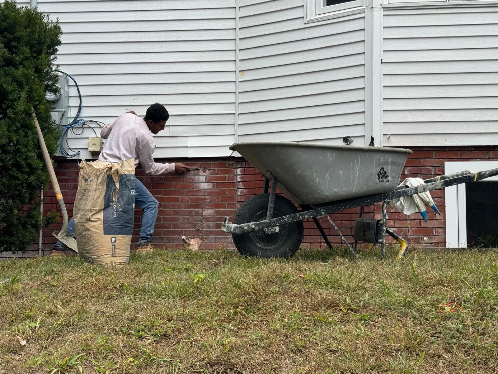 A person works on repairing the brick foundation of a house, using a trowel next to a wheelbarrow and a bag of mortar.