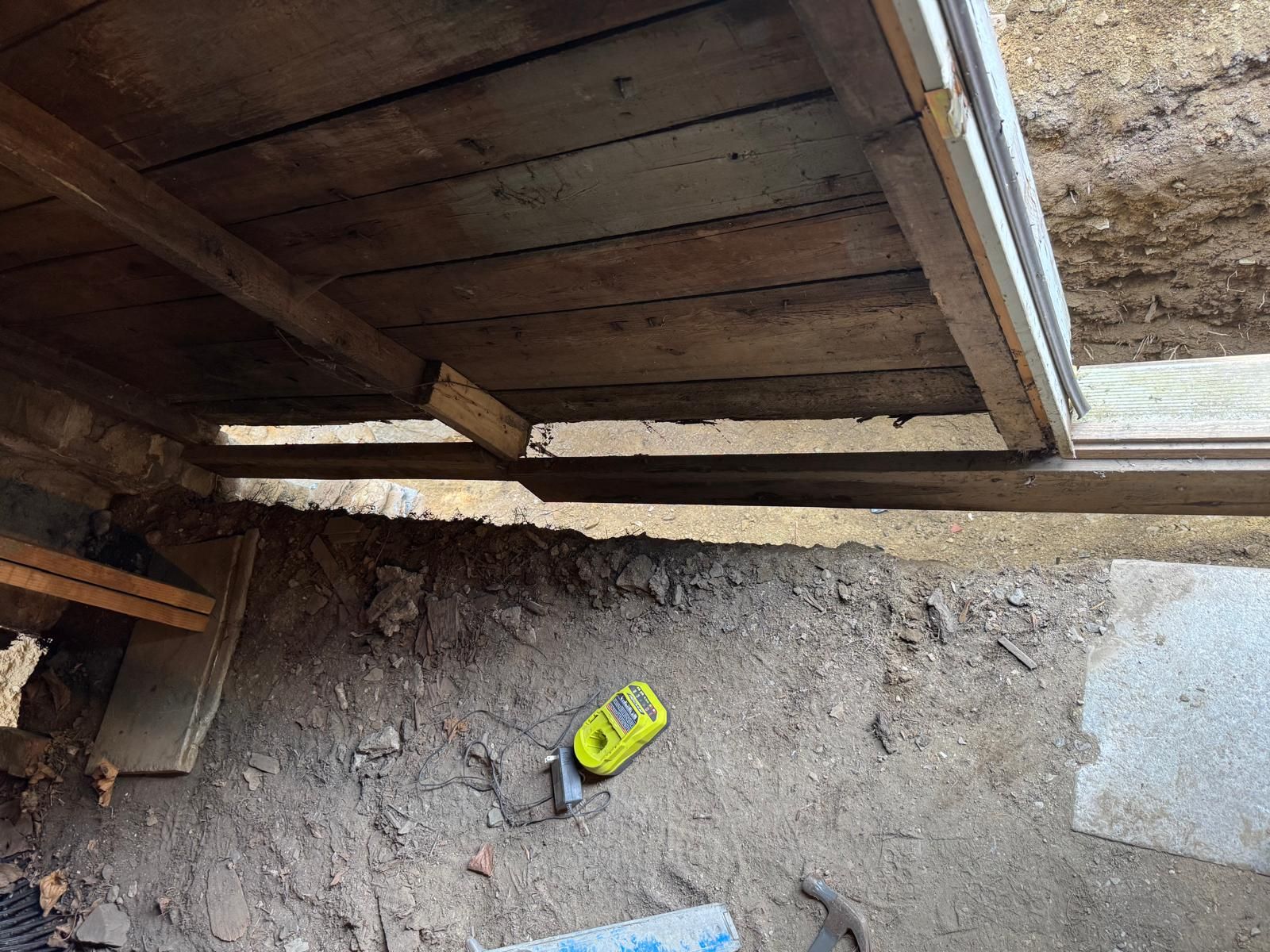 A view from inside a structure showing a wooden wall frame with a gap near the ground, revealing soil and a yellow tool.