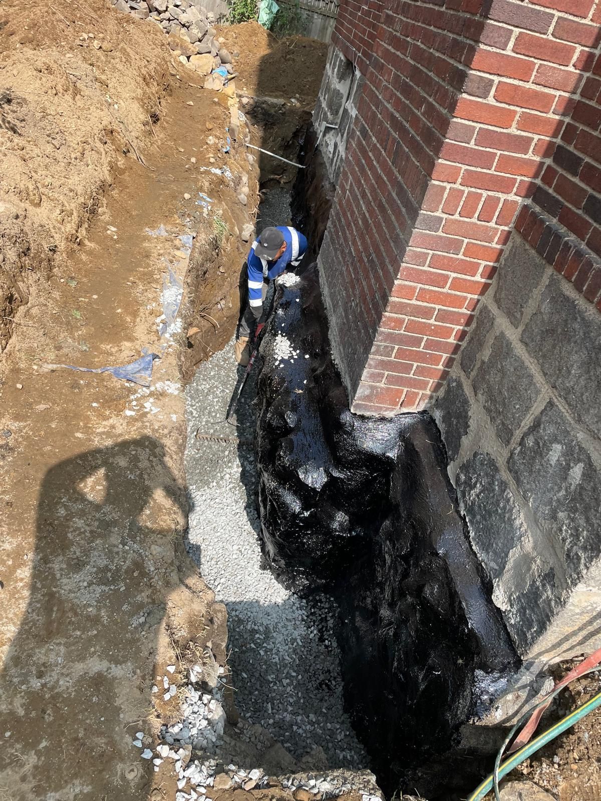 A worker kneeling in a trench beside a brick building foundation that has been coated with black waterproofing material.