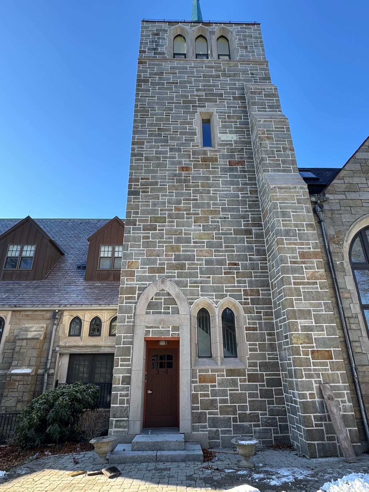 A tall stone bell tower attached to a stone church building under a clear blue sky, with a brown arched wooden door.