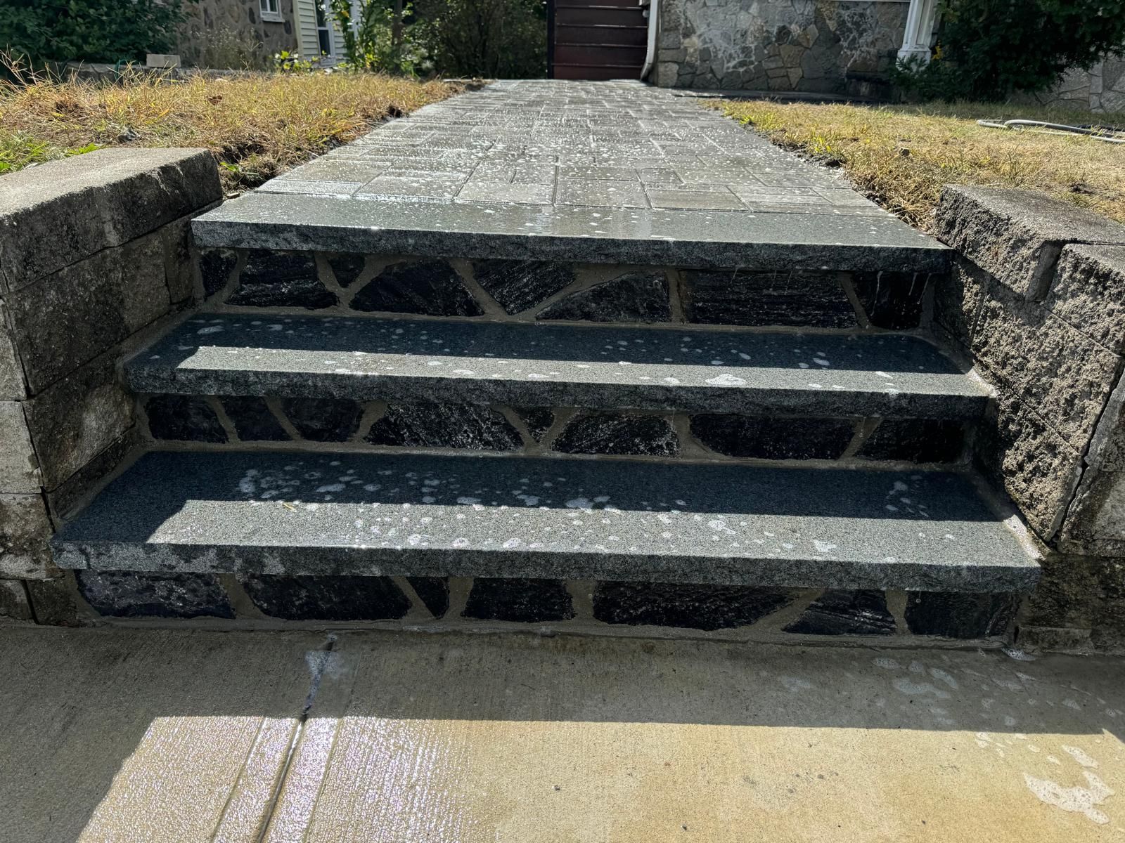 A set of three dark stone steps with a geometric mortar pattern leading to a paved walkway outdoors.