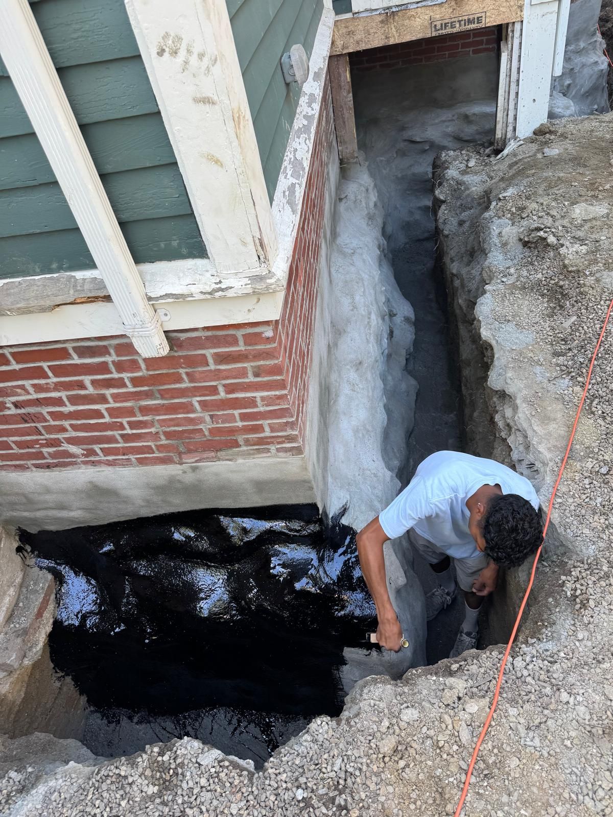 A person applies black waterproof sealant to the foundation of a brick house in an excavated trench.