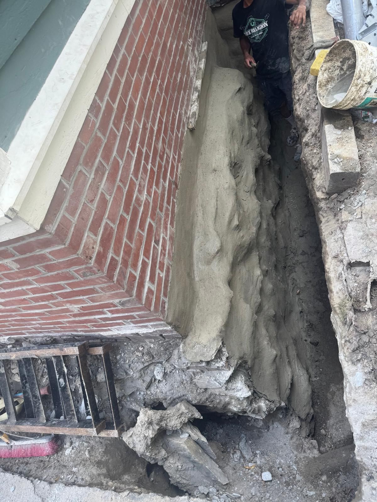 A trench dug along a brick building foundation, showing a worker applying cement coating to the wall surface.
