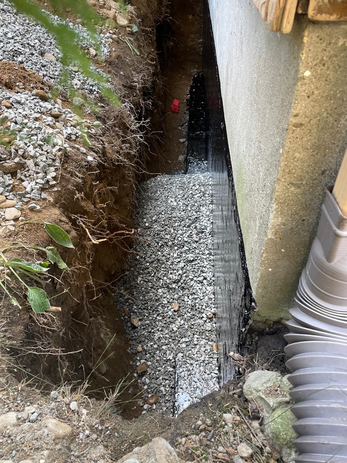 A narrow, excavated trench along a building foundation featuring a black drainage membrane and gravel backfill.