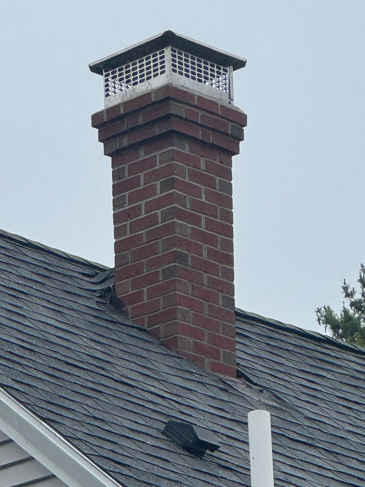 A red brick chimney rises from a shingled roof, topped with a metal flue cover against a cloudy sky.