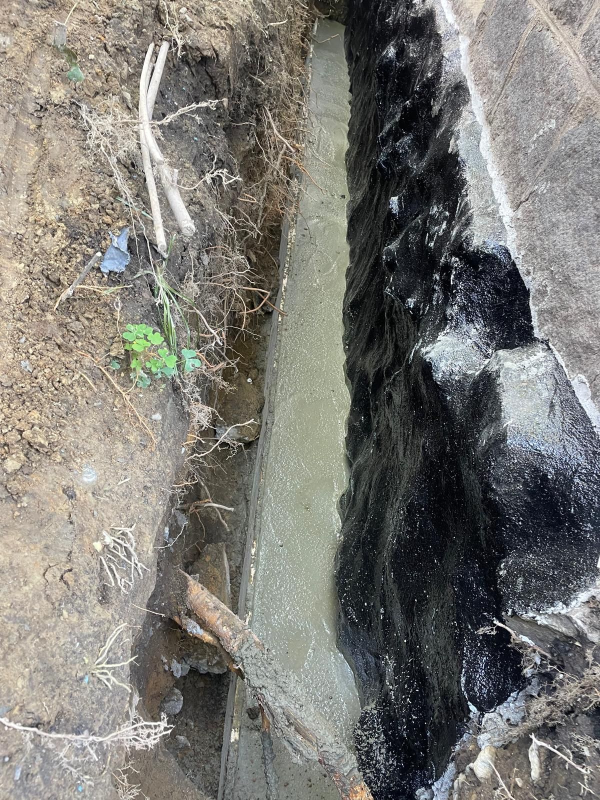 A narrow trench beside a foundation wall coated in black waterproof sealant, with a fresh concrete footer at the bottom.